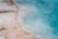 A close-up view of a geothermal hot spring, featuring mineral deposits and colorful patterns. The transition between the rocky edges and the clear, turquoise water creates a striking contrast. The water appears misty due to steam rising from the surface.