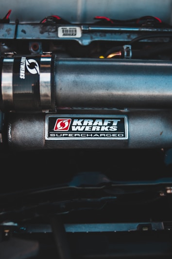 A close-up of a car engine featuring a metallic component with a 'Kraft Werks Supercharged' label visible. The setup includes other engine parts and piping, all housed in a compartment with a clean, industrial look. Wiring is seen at the top section, and the main color of the engine parts is gray, with some labels in red and white.
