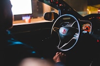 A person is sitting inside a car with a focus on the Honda steering wheel and illuminated dashboard. The scene is dimly lit, giving a cozy and slightly mysterious ambiance, suggesting it could be nighttime. Reflections and lights from the outside world can be seen through the car windows.