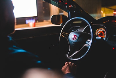 A person is sitting inside a car with a focus on the Honda steering wheel and illuminated dashboard. The scene is dimly lit, giving a cozy and slightly mysterious ambiance, suggesting it could be nighttime. Reflections and lights from the outside world can be seen through the car windows.