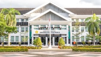 The image depicts a large, white building with an architectural style featuring prominent gabled roofs and columns at the entrance. The facade includes several glass windows, and there is a sign in the center with the words 'Universitas Syiah Kuala.' In front of the building are neatly arranged plants and trees, including palm trees, creating a landscaped garden area. A flagpole with a flag flying is also visible.
