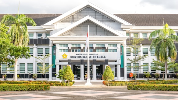 The image depicts a large, white building with an architectural style featuring prominent gabled roofs and columns at the entrance. The facade includes several glass windows, and there is a sign in the center with the words 'Universitas Syiah Kuala.' In front of the building are neatly arranged plants and trees, including palm trees, creating a landscaped garden area. A flagpole with a flag flying is also visible.