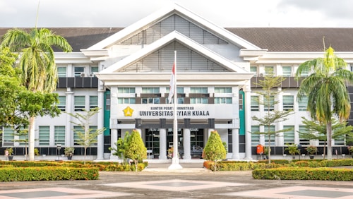 The image depicts a large, white building with an architectural style featuring prominent gabled roofs and columns at the entrance. The facade includes several glass windows, and there is a sign in the center with the words 'Universitas Syiah Kuala.' In front of the building are neatly arranged plants and trees, including palm trees, creating a landscaped garden area. A flagpole with a flag flying is also visible.