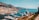 A coastal city with a bustling marina filled with numerous yachts and small boats. Tall buildings and hotels rise up along the shore, surrounded by mountainous terrain in the background. The ocean is calm and reflects the clear blue sky, creating a picturesque and luxurious atmosphere.