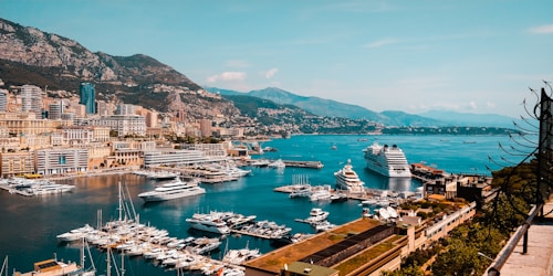 A coastal city with a bustling marina filled with numerous yachts and small boats. Tall buildings and hotels rise up along the shore, surrounded by mountainous terrain in the background. The ocean is calm and reflects the clear blue sky, creating a picturesque and luxurious atmosphere.