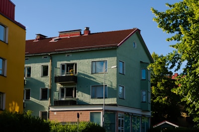 A two-story residential building with green exterior walls and a dark red sloped roof. The building features several windows and small balconies, with a single streetlamp in front. The sun casts long shadows, indicating late afternoon sunlight. Trees with lush green leaves are visible in the background, suggesting a suburban neighborhood setting.