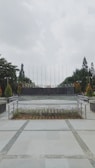 A university campus area with a paved walkway leading to a circular plaza surrounded by neatly arranged plants. In the background, large letters spell out 'UNIVERSITAS,' flanked by trees and tall flagpoles. Overhead, the sky is overcast, creating a calm and serene atmosphere.