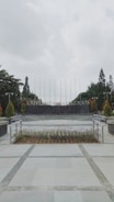 A university campus area with a paved walkway leading to a circular plaza surrounded by neatly arranged plants. In the background, large letters spell out 'UNIVERSITAS,' flanked by trees and tall flagpoles. Overhead, the sky is overcast, creating a calm and serene atmosphere.