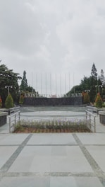 A university campus area with a paved walkway leading to a circular plaza surrounded by neatly arranged plants. In the background, large letters spell out 'UNIVERSITAS,' flanked by trees and tall flagpoles. Overhead, the sky is overcast, creating a calm and serene atmosphere.