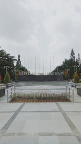 A university campus area with a paved walkway leading to a circular plaza surrounded by neatly arranged plants. In the background, large letters spell out 'UNIVERSITAS,' flanked by trees and tall flagpoles. Overhead, the sky is overcast, creating a calm and serene atmosphere.