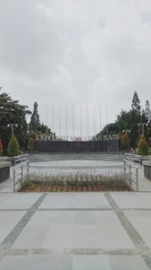 A university campus area with a paved walkway leading to a circular plaza surrounded by neatly arranged plants. In the background, large letters spell out 'UNIVERSITAS,' flanked by trees and tall flagpoles. Overhead, the sky is overcast, creating a calm and serene atmosphere.
