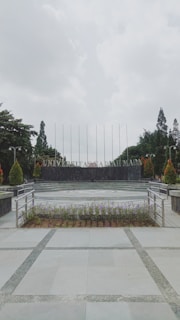 A university campus area with a paved walkway leading to a circular plaza surrounded by neatly arranged plants. In the background, large letters spell out 'UNIVERSITAS,' flanked by trees and tall flagpoles. Overhead, the sky is overcast, creating a calm and serene atmosphere.