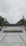 A university campus area with a paved walkway leading to a circular plaza surrounded by neatly arranged plants. In the background, large letters spell out 'UNIVERSITAS,' flanked by trees and tall flagpoles. Overhead, the sky is overcast, creating a calm and serene atmosphere.