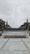 A university campus area with a paved walkway leading to a circular plaza surrounded by neatly arranged plants. In the background, large letters spell out 'UNIVERSITAS,' flanked by trees and tall flagpoles. Overhead, the sky is overcast, creating a calm and serene atmosphere.