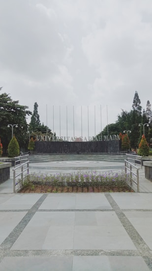 A university campus area with a paved walkway leading to a circular plaza surrounded by neatly arranged plants. In the background, large letters spell out 'UNIVERSITAS,' flanked by trees and tall flagpoles. Overhead, the sky is overcast, creating a calm and serene atmosphere.