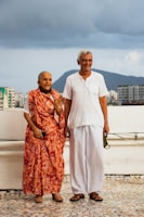 An elderly couple stands on a tiled surface outdoors, with a cloudy sky and a cityscape in the background. The woman is wearing a vibrant orange sari with floral patterns, while the man wears a simple white shirt and pants. Both are smiling and appear to be enjoying a moment together.