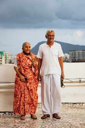 An elderly couple stands on a tiled surface outdoors, with a cloudy sky and a cityscape in the background. The woman is wearing a vibrant orange sari with floral patterns, while the man wears a simple white shirt and pants. Both are smiling and appear to be enjoying a moment together.