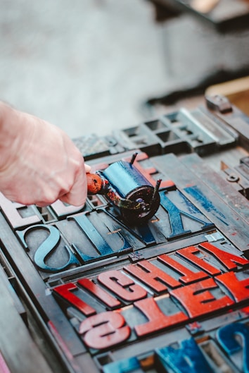 A hand is applying ink with a roller onto wooden or metal type blocks arranged for letterpress printing. The blocks display a variety of letters in different sizes and orientations, some of which are inked in red and blue.