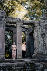 Two people dressed in traditional clothing stand near ancient stone ruins surrounded by lush greenery. The structure features tall, weathered columns and intricate carvings, suggesting historical significance.