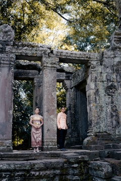Two people dressed in traditional clothing stand near ancient stone ruins surrounded by lush greenery. The structure features tall, weathered columns and intricate carvings, suggesting historical significance.