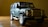 A toy model of a Land Rover vehicle is placed on a wooden surface. The model is detailed, featuring a dark green body, prominent front grille, and realistic-looking tires. The lighting casts shadows that accentuate the textures and details of the model.