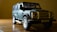A toy model of a Land Rover vehicle is placed on a wooden surface. The model is detailed, featuring a dark green body, prominent front grille, and realistic-looking tires. The lighting casts shadows that accentuate the textures and details of the model.
