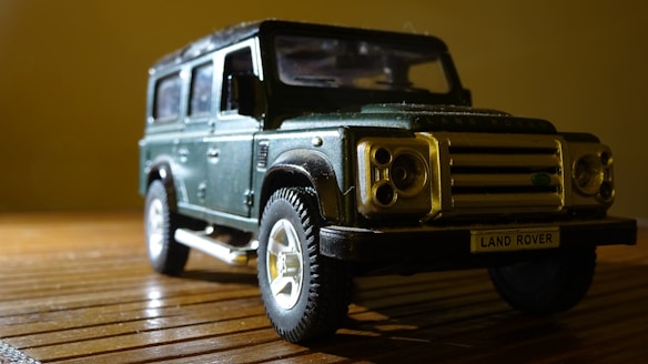 A toy model of a Land Rover vehicle is placed on a wooden surface. The model is detailed, featuring a dark green body, prominent front grille, and realistic-looking tires. The lighting casts shadows that accentuate the textures and details of the model.