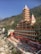A tall, multi-tiered Hindu temple with red and white architecture stands prominently against a backdrop of lush green hills. The building's intricate design includes several small spires and decorative elements. The ground level features a courtyard with various pathways and steps leading down to a rocky riverbank. Clear blue skies are visible above.