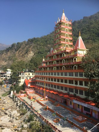 A tall, multi-tiered Hindu temple with red and white architecture stands prominently against a backdrop of lush green hills. The building's intricate design includes several small spires and decorative elements. The ground level features a courtyard with various pathways and steps leading down to a rocky riverbank. Clear blue skies are visible above.