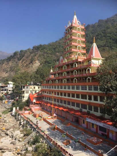 A tall, multi-tiered Hindu temple with red and white architecture stands prominently against a backdrop of lush green hills. The building's intricate design includes several small spires and decorative elements. The ground level features a courtyard with various pathways and steps leading down to a rocky riverbank. Clear blue skies are visible above.
