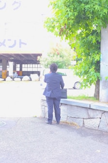 A person wearing a dark suit is standing on a sidewalk facing away and holding a briefcase. There is a stone ledge beside them, and the background includes greenery on the right and a lightly trafficked urban area with a white vehicle in the distance.