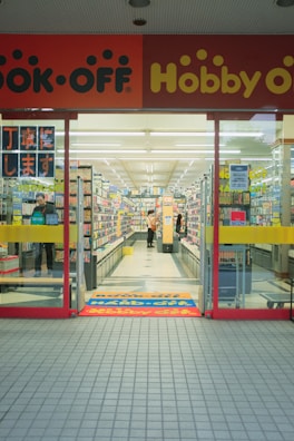 The entrance of a store, possibly a secondhand book or hobby shop. The interior is brightly lit, with rows of shelves filled with various items. Two people are visible inside, browsing or shopping. The signage above the entrance is colorful, mainly in red and yellow, and displays the store's name prominently.