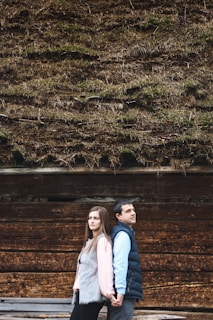 A couple standing back-to-back, holding hands in front of a wooden wall with a thatched roof covered in moss. The woman on the left is wearing a pink top and gray fur vest, while the man on the right is dressed in a blue shirt and dark vest.