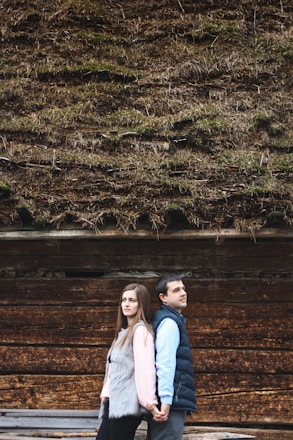 A couple standing back-to-back, holding hands in front of a wooden wall with a thatched roof covered in moss. The woman on the left is wearing a pink top and gray fur vest, while the man on the right is dressed in a blue shirt and dark vest.