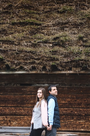 A couple standing back-to-back, holding hands in front of a wooden wall with a thatched roof covered in moss. The woman on the left is wearing a pink top and gray fur vest, while the man on the right is dressed in a blue shirt and dark vest.
