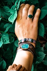 A hand adorned with a green gemstone ring and beaded bracelet resting on lush green leaves. The wrist features a leather strap bracelet with a decorative turquoise disc.