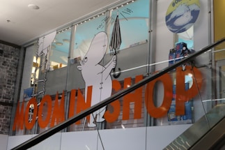 A store display featuring cartoon characters with a focus on a white character holding an umbrella. Large orange letters spell out 'MOOMIN SHOP' across the glass. The background consists of a retail interior and another cartoon in a circle with hashtags.