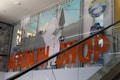 A store display featuring cartoon characters with a focus on a white character holding an umbrella. Large orange letters spell out 'MOOMIN SHOP' across the glass. The background consists of a retail interior and another cartoon in a circle with hashtags.