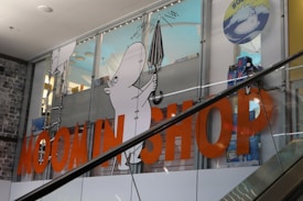 A store display featuring cartoon characters with a focus on a white character holding an umbrella. Large orange letters spell out 'MOOMIN SHOP' across the glass. The background consists of a retail interior and another cartoon in a circle with hashtags.