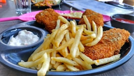 A plate filled with golden french fries and two pieces of crispy fried fish. The dish is accompanied by a small serving of tartar sauce on the left and a dipping sauce on the right. The setting is casual with a pink fork placed beside the plate and a glass with a drink in the background.