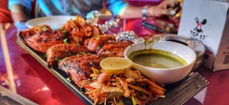 A plate of grilled chicken pieces seasoned with herbs is accompanied by a bowl of green chutney and a serving of fresh salad with onions and lemon on top. In the background, a person is seated at a table with a pink tablecloth and a menu card with a restaurant logo.