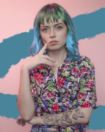 A person with multicolored hair including shades of green and blue, wearing a vibrant floral shirt. They have a thoughtful expression, with their finger resting on their face. Visible tattoos on their arm depict a butterfly and dice among other designs. The background is a mix of pink and teal with an abstract pattern.