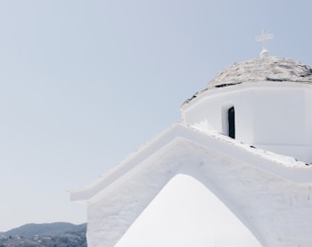 A whitewashed church with a domed roof and a cross on top is set against a clear blue sky. The architecture features simple, clean lines and is typical of Greek island designs. Rolling hills with vegetation are visible in the background.