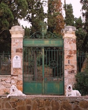 Two white dogs are resting in front of an ornate green gate made of metal. The gate is flanked by stone pillars and leads into a wooded area. A sign with text and a cross symbol is affixed to one of the pillars. The background is filled with tall trees providing a natural setting.