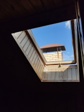 A view through a skylight showing a brick chimney with a red metal roof. The blue sky and some clouds can be seen beyond the chimney. The interior framing of the skylight is made of light-colored wood.