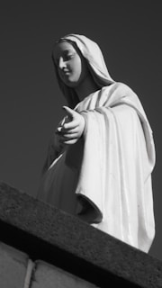 A statue of a figure in robes with an outstretched hand, appearing serene and composed, is set against a deep, monochromatic background. The figure's facial expression is calm and peaceful.