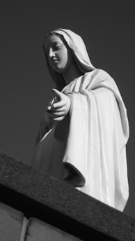 A statue of a figure in robes with an outstretched hand, appearing serene and composed, is set against a deep, monochromatic background. The figure's facial expression is calm and peaceful.