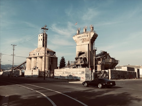 An industrial scene with large concrete silos and a processing plant. The structures have graffiti on the walls and the words 'Walkers Concrete Inc' can be seen on one building. A vintage car is parked on the road in the foreground.