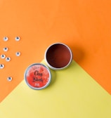 A container of clay blush with a labeled lid is open on a bright, two-toned orange and yellow background. Several small, shiny gems are scattered nearby.