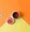 A container of clay blush with a labeled lid is open on a bright, two-toned orange and yellow background. Several small, shiny gems are scattered nearby.